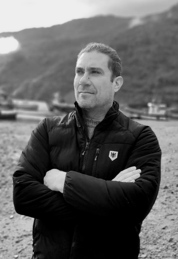 Retrato en blanco y negro de un hombre con brazos cruzados, luciendo una chaqueta negra, en un entorno al aire libre con montañas al fondo.